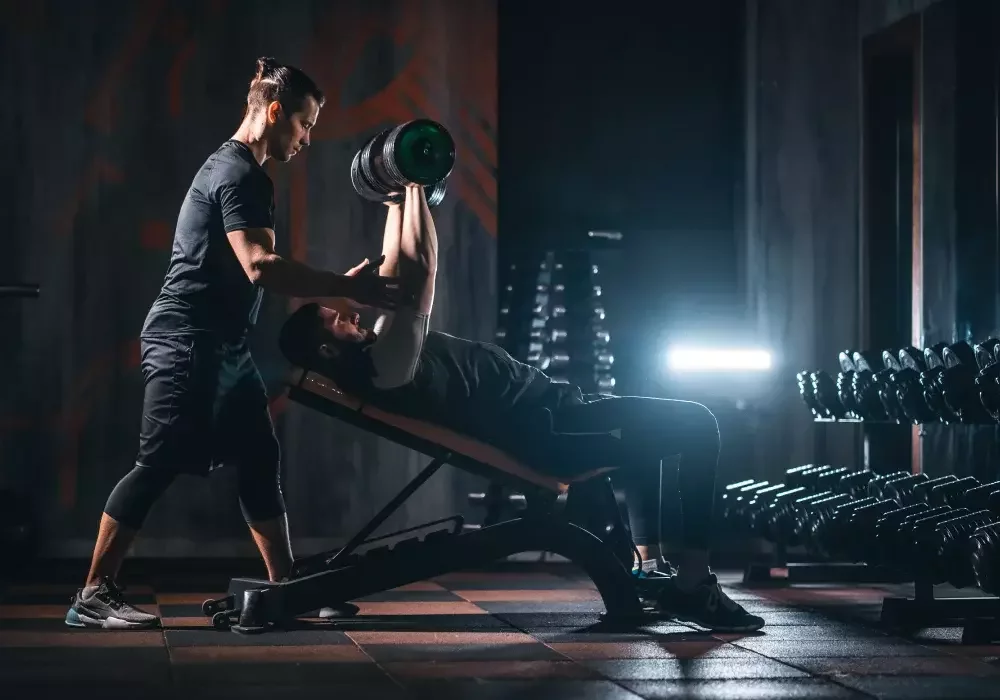 A spotter spotting a man raising dumbbells over his head on a bench at the gym.