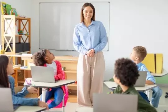 A teacher stands at the back of a clean classroom and addresses children sitting at their desks with laptops.