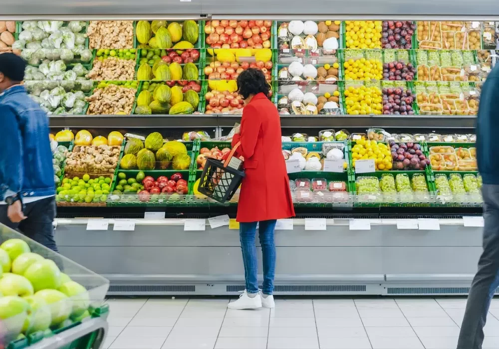 A woman in a red coat shops for fruit at a supermarket aisle.